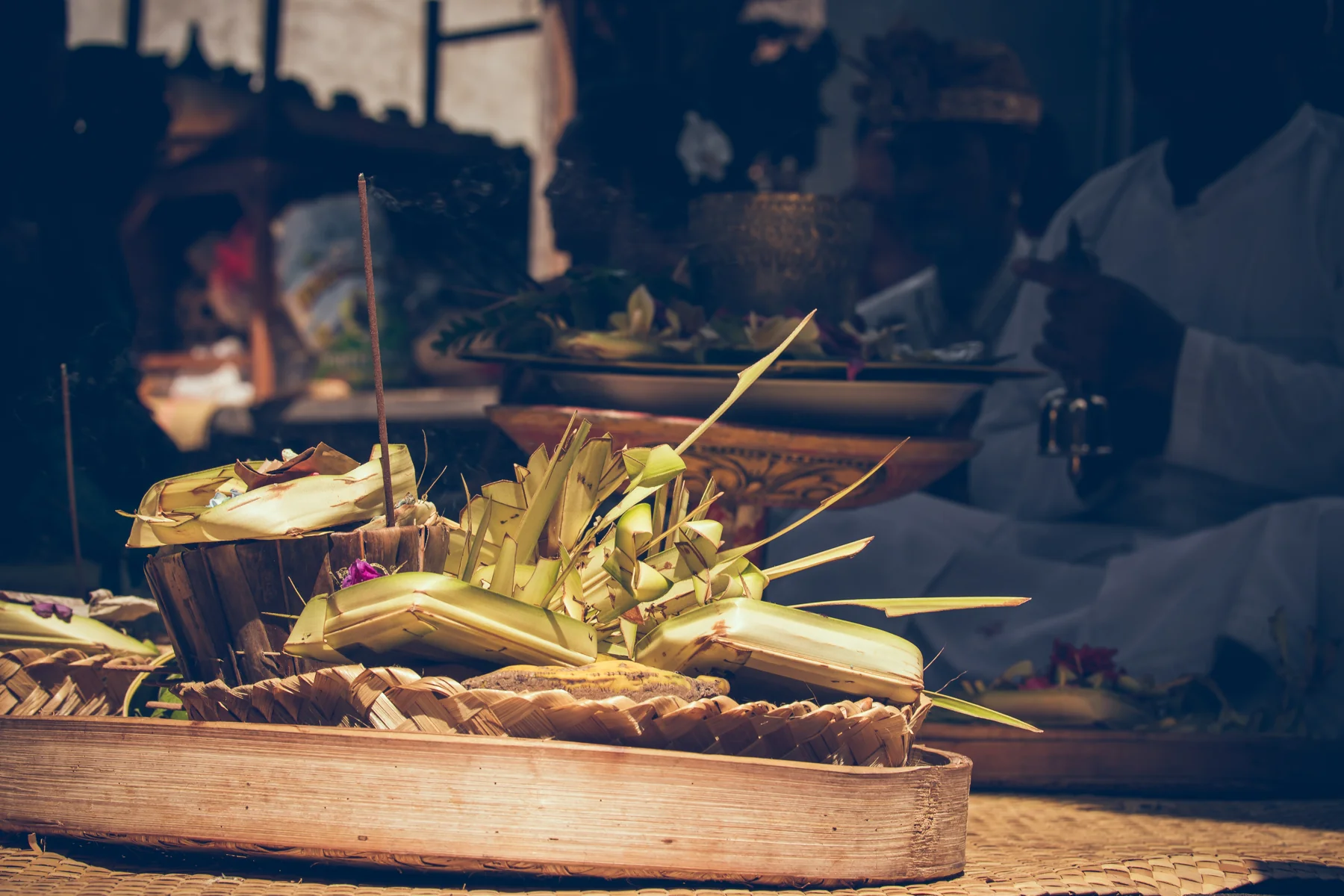 Palm-leaf offerings and incense sit on a wooden work surface with artisans behind them.