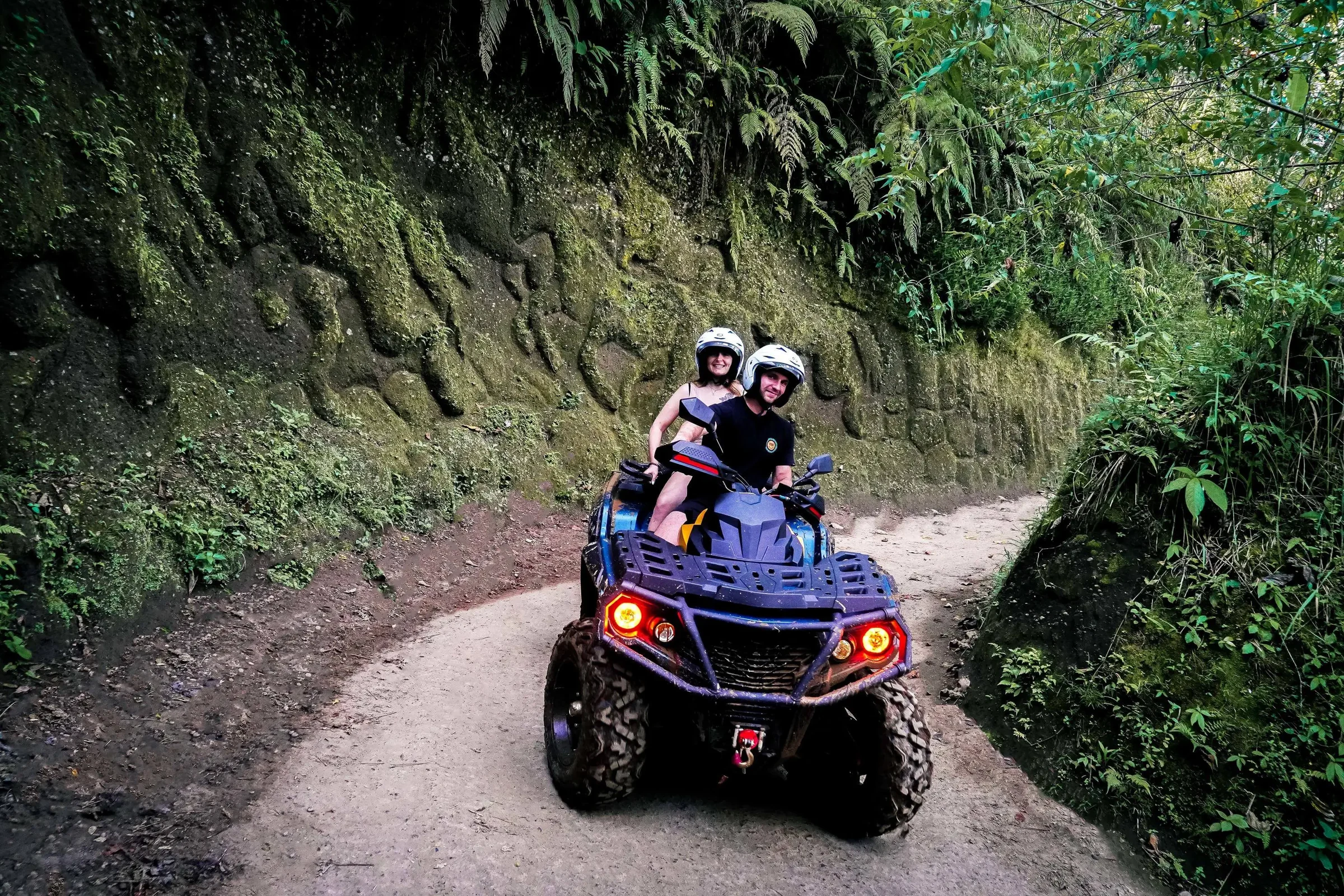 Two helmeted riders on a tandem ATV follow a tree-lined trail at Alasan Adventure