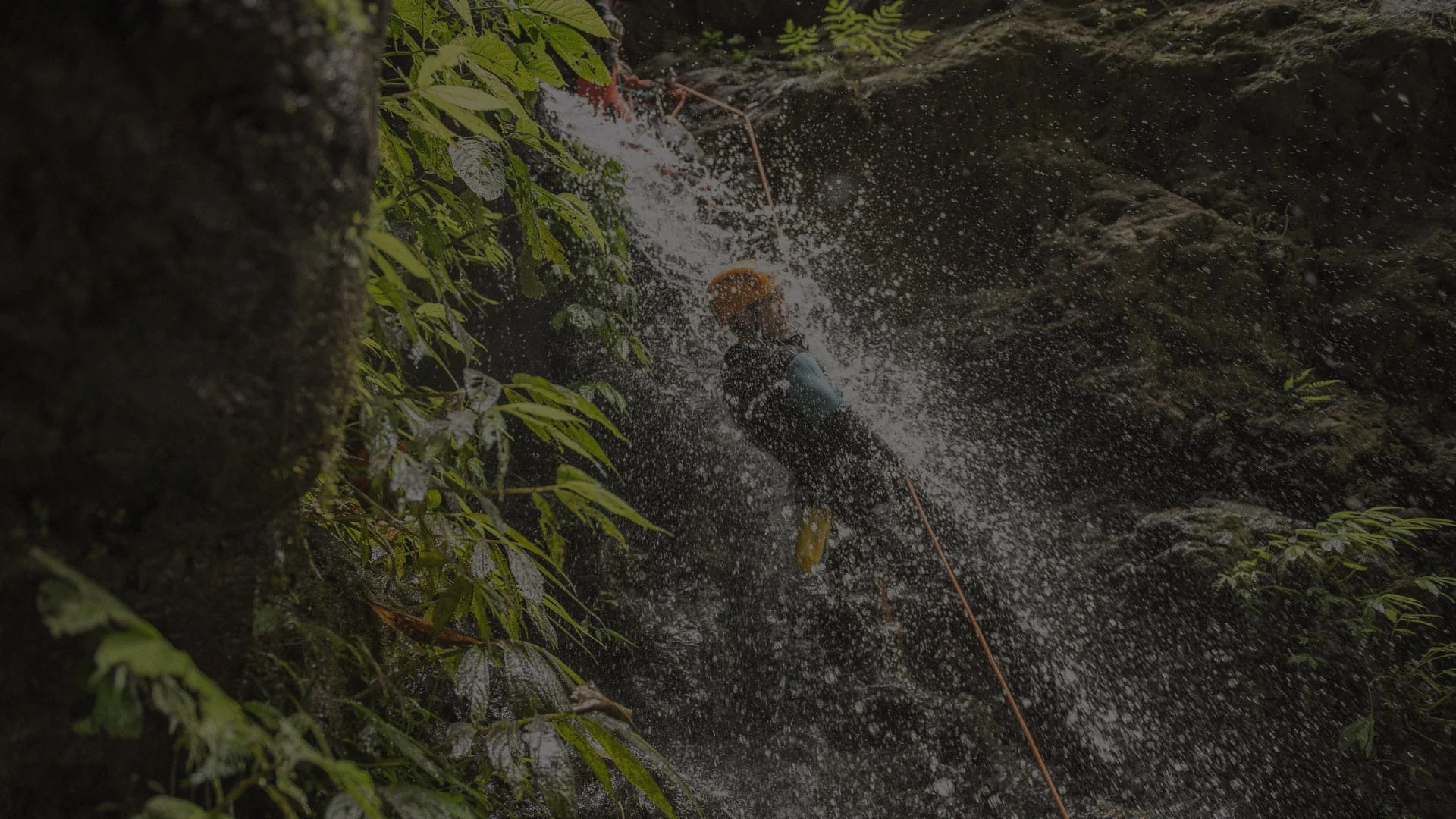 A helmeted canyoner rappels through a waterfall between mossy volcanic rock walls in Kalimudah