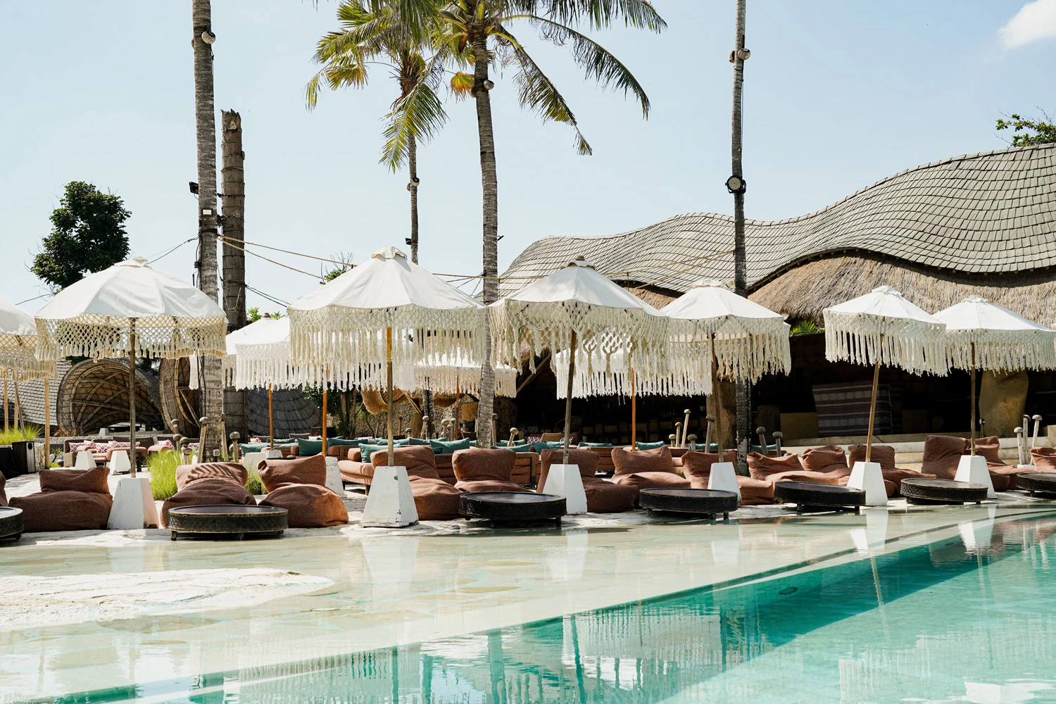 Mari Beach Club pool with fringed white parasols, palms and curved thatched roof.