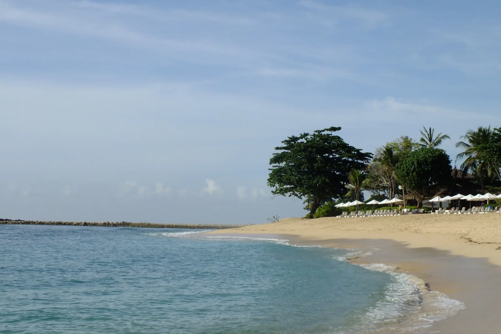 Calm blue water meeting the sandy Nusa Dua shoreline in Bali, with trees beside the beach.