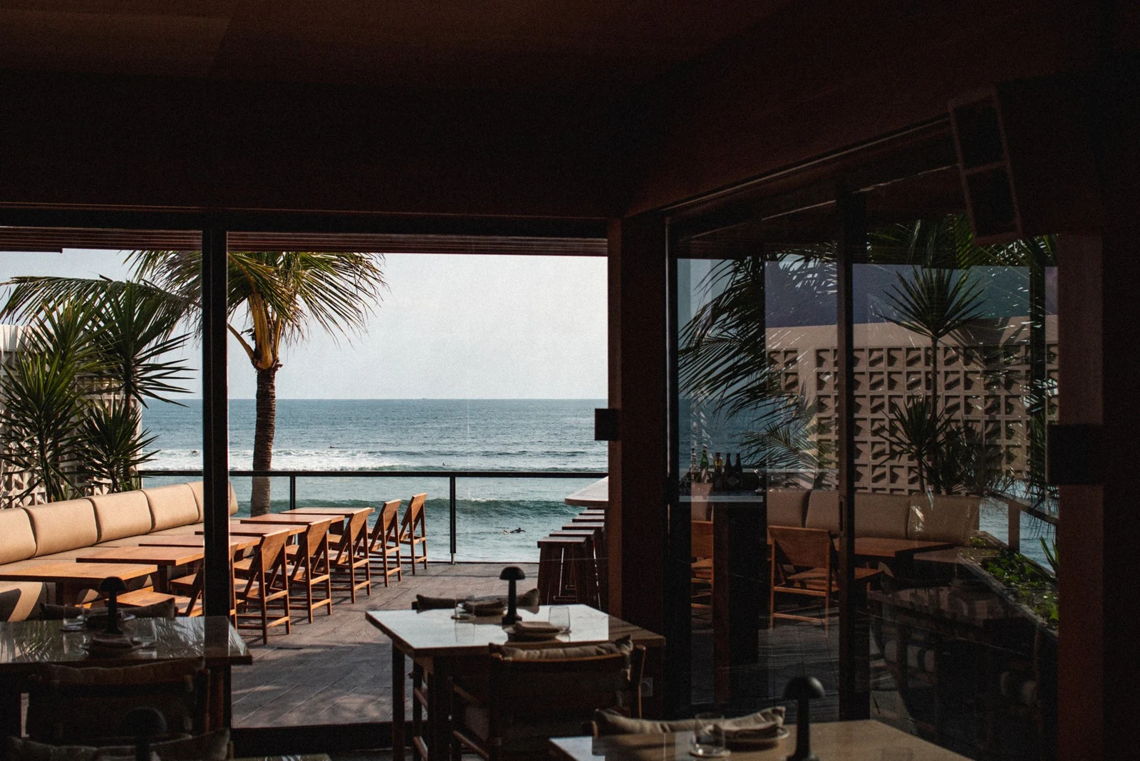 Ocean-facing tables and open timber-framed windows at SKOOL Kitchen in soft evening light.