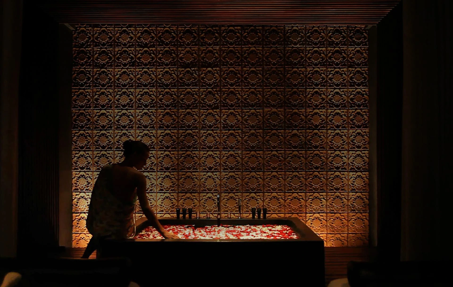 A spa attendant prepares a flower-filled bath against an illuminated carved wall at Soori Spa.