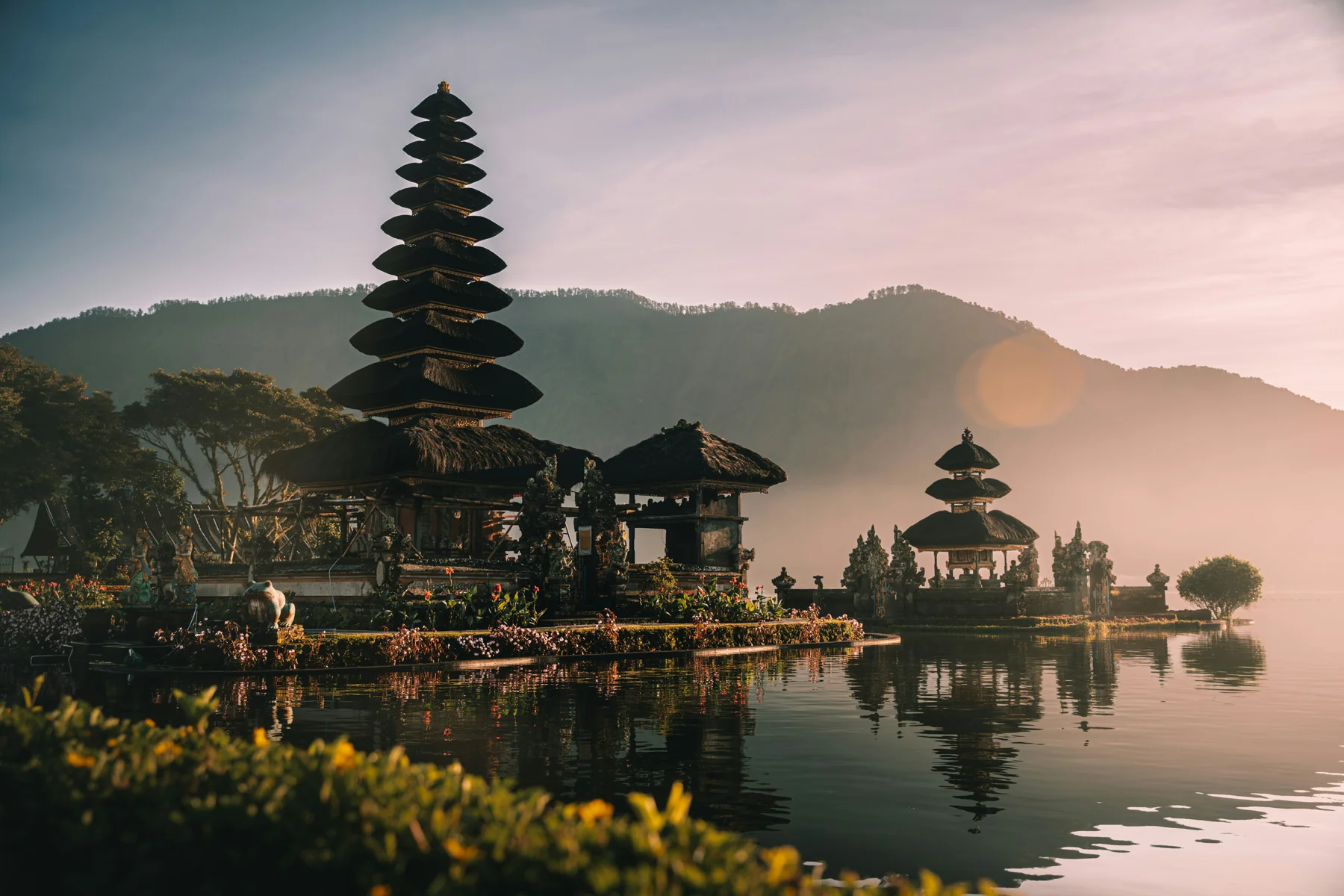 Tiered temple shrines at Ulun Danu Beratan stand beside still lake water and hazy mountain slopes.