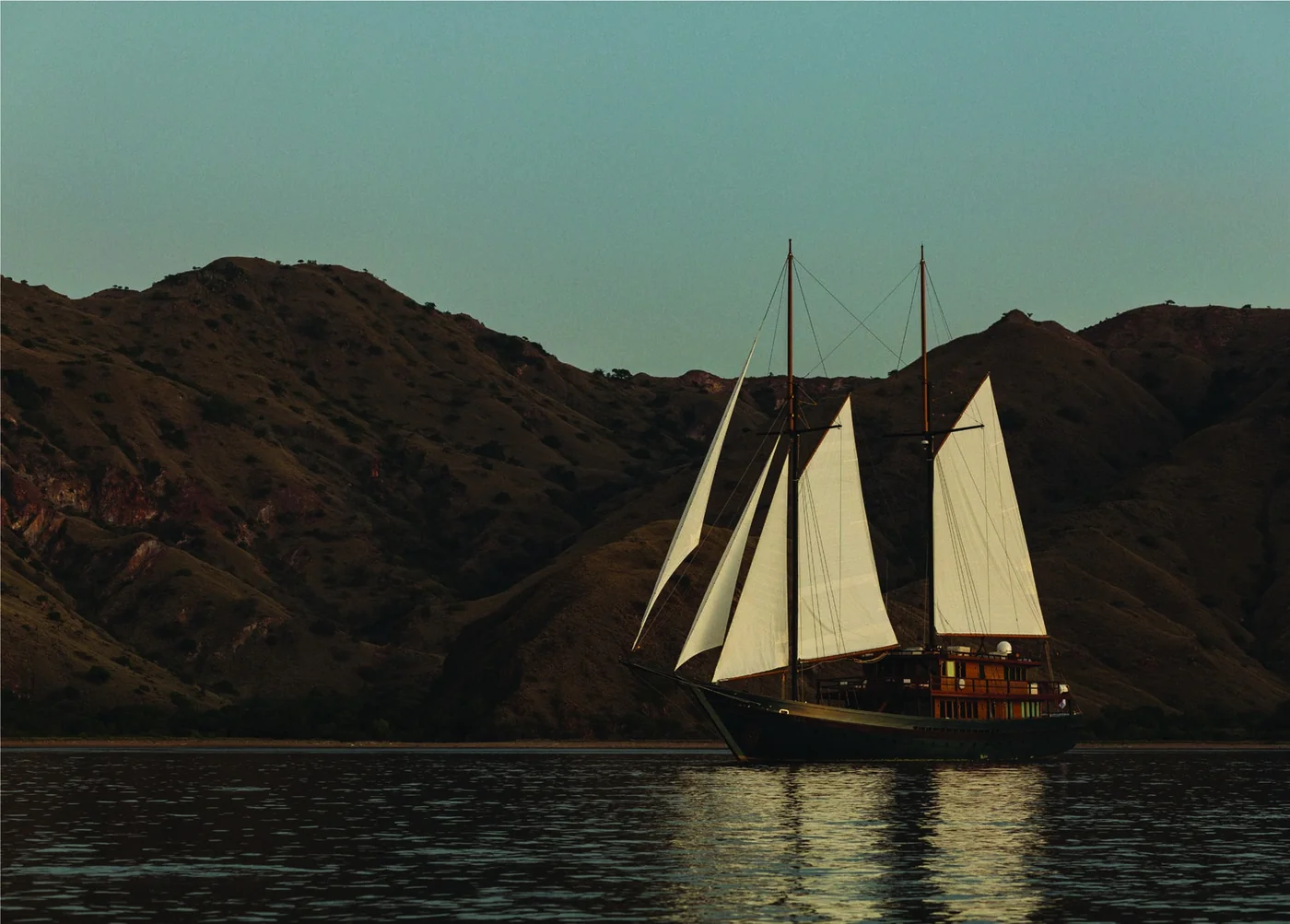 Vela sailing yacht at dusk with sails raised beside rugged island hills